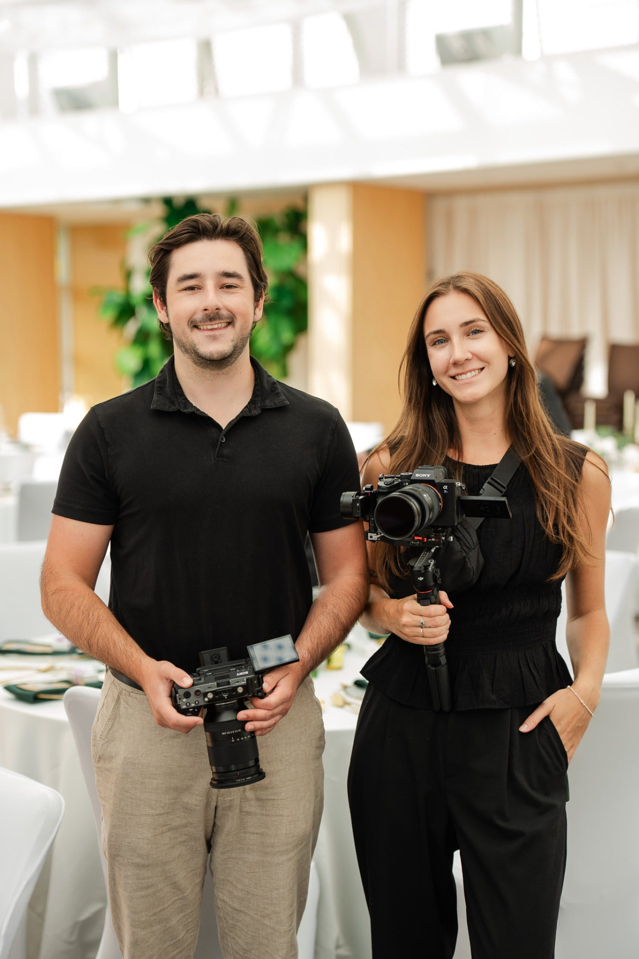 Two videographers, a man and a woman, holding professional cameras in a bright white event space